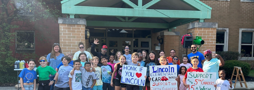 A group of students, teachers and parents gathered in front of a school at a car wash.